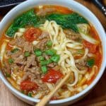 Tomato beef noodle soup with garnishes, thick noodles, and beef in a white bowl