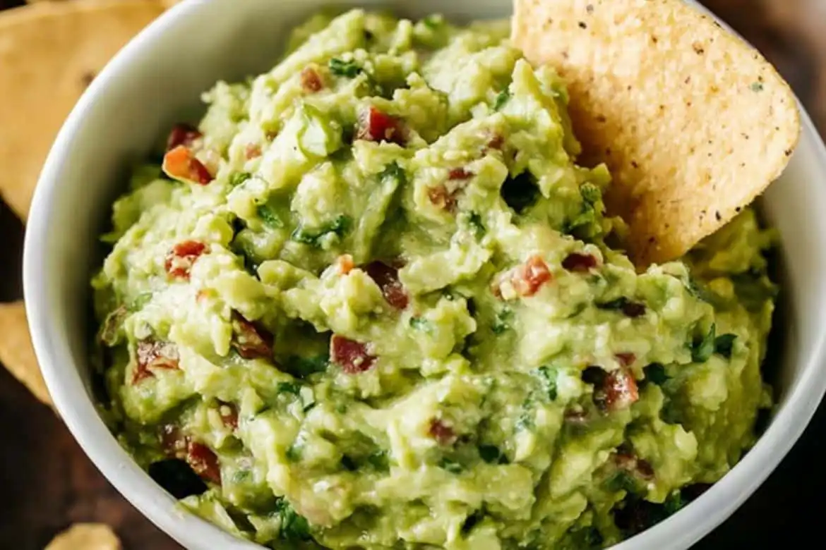 Guacamole recipe made with ripe avocados, lime, cilantro, and tomatoes in a white bowl with tortilla chip