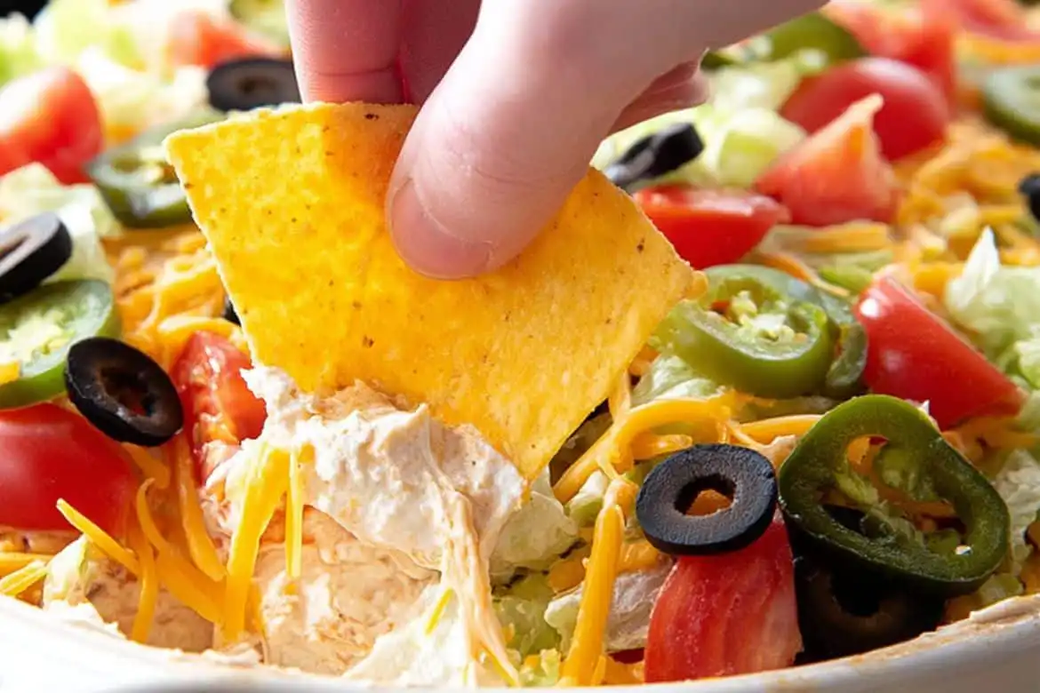 Hand dipping tortilla chip into creamy taco dip with cream cheese, topped with lettuce, olives, jalapeños, and tomatoes