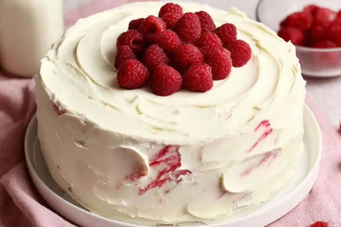 White chocolate raspberry cake with creamy swirled frosting and fresh raspberries on top on a white plate
